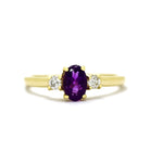 A product photo of a yellow gold amethyst and diamond trio ring sitting on a white background. The 6x4mm oval purple amethyst gemstone stands in stark contrast to the 0.03ct white diamond stones on either side.