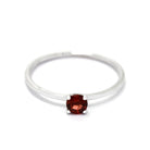 A product photo of a delicate white gold stacking ring with a tiny, claw-set garnet in the centre sitting on a white background. The band is slim and thread-like, with the focus drawn to the petite 4mm glinting garnet gemstone.