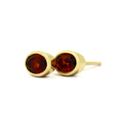 A product photo of a pair of garnet earrings in 9ct yellow gold sitting on a white background. The fine 6x4mm garnet jewels are framed in a thick layer of yellow gold in a bezel setting.