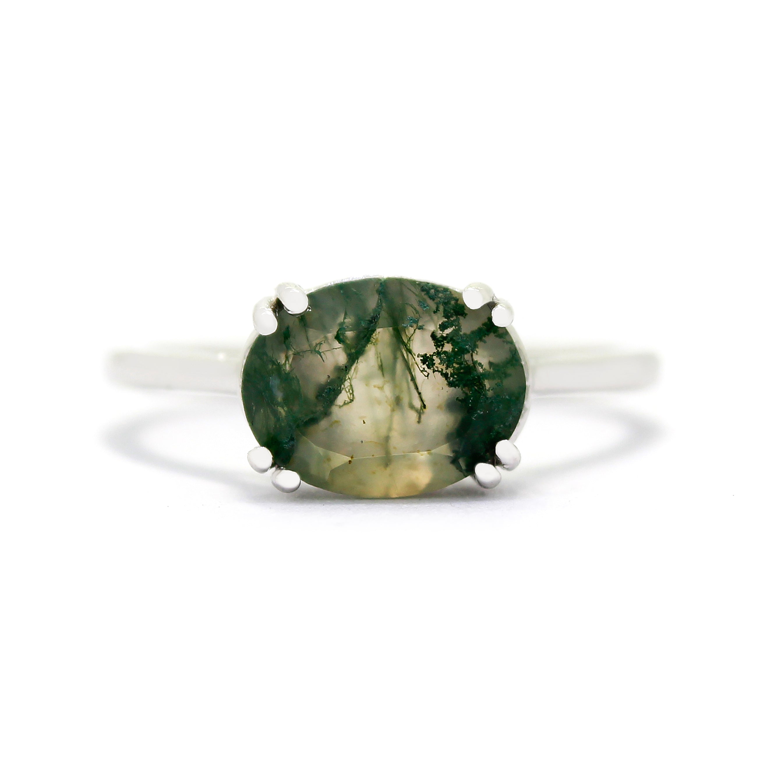 A product photo of a 9 karat white gold ring with an impressive oval moss agate centre stone sitting on a white background. The band is simple and rounded gold, with a single 10x8mm moss agate centre stone - adorned with moss-like, swirling dendritic inclusions. The moss agate stone is semi-opaque light olive colour, with striking mossy inclusions throughout.