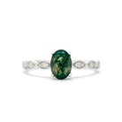 A product photo of an ornate moss agate and diamond ring in 9k white gold. Oval gold frames, adorned with two small diamond gems each, pattern the band before smoothing out towards the back. In the centre sits a deeply-included, dark green moss agate jewel held in place by 4 golden claws.