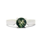 A product photo of a moss agate solitaire ring in 9 karat white gold sitting on a white background. The smooth golden band has a slightly different shape to the classic solitaire look - with flat, tall edges tapering to thin points on either side of the round, semi-opaque moss agate gemstone.