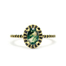 A product photo of a moss agate ring in 9k yellow gold on a white background. A large, brilliant oval jewel in the centre reflects swirling, ink-like dendritic inclusions, and is framed by a ring of black diamonds. A simple round band continues the diamond detailing for half of its length, smoothing out into a simple yellow gold back. The moss agate stone is semi-opaque white, with striking mossy inclusions and tendrils throughout.