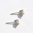 A product photo of a pair of 9 karat white gold lab diamond stud earrings sitting in the sun on a white textured background. The brilliant, colourless half pointer round diamonds measure 5mm across, and are held in place by a simple 4-claw setting.