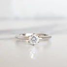 A product photo of a 14 karat white gold lab diamond solitaire engagement ring sitting in the sun on a white textured background. The brilliant, colourless half pointer round diamond measures 5mm across, and is held in place by 6 delicate claws in a high-profile prong setting.