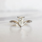 A product photo of a 14 karat white gold lab diamond solitaire engagement ring sitting in the sun on a white textured background. The brilliant, colourless 0.70ct pear-shaped diamond measures around 7mm lengthwise and is held in place by 3 delicate claws, nestled in the base of a curved gold band that tapers into a sharp point at the narrowest part of the pear in a low-profile prong setting.