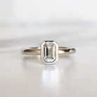 A product photo of a 14 karat white gold lab diamond bezel solitaire engagement ring sitting in the sun on a white textured background. The brilliant, colourless 0.72ct emerald-cut diamond measures about 6.3mm lengthwise, and is held in place by a delicate golden-framed bezel setting.