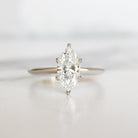 A product photo of a 14 karat white gold lab diamond solitaire engagement ring sitting in the sun on a white textured background. The brilliant, colourless 1 carat marquise-cut diamond measures 10.7mm lengthwise, and is held in place by 6 delicate claws in a high-profile prong setting.