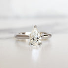 A product photo of a 14 karat white gold lab diamond solitaire engagement ring sitting in the sun on a white textured background. The brilliant, colourless 1 carat pear-cut diamond measures 8.73mm lengthwise, and is held in place by 3 delicate claws in a high-profile cathedral setting in the middle of a tapered band.