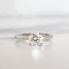 A product photo of a 14 karat white gold lab diamond solitaire engagement ring sitting in the sun on a white textured background. The brilliant, colourless 1 carat round-cut diamond measures 6.3mm lengthwise, and is held in place by 4 delicate claws in a prong setting.