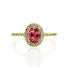 A product photo of a pink tourmaline ring in 9 karat yellow gold on a white background. A large, brilliant oval jewel in the centre reflects warm pink-toned light, and is framed by a halo ring of white diamonds. A simple round band continues the diamond detailing for half of its length, smoothing out into a simple yellow gold back.