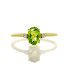 A product photo of a yellow gold peridot ring sitting against a white background. The yellow gold band is plain and smooth, and the centre oval-cut grass green peridot stone is framed by a single white diamond on either side.