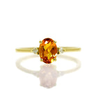 A product photo of a yellow gold citrine ring sitting against a white background. The yellow gold band is plain and smooth, and the centre oval-cut bright orange citrine stone is framed by a single white diamond on either side.