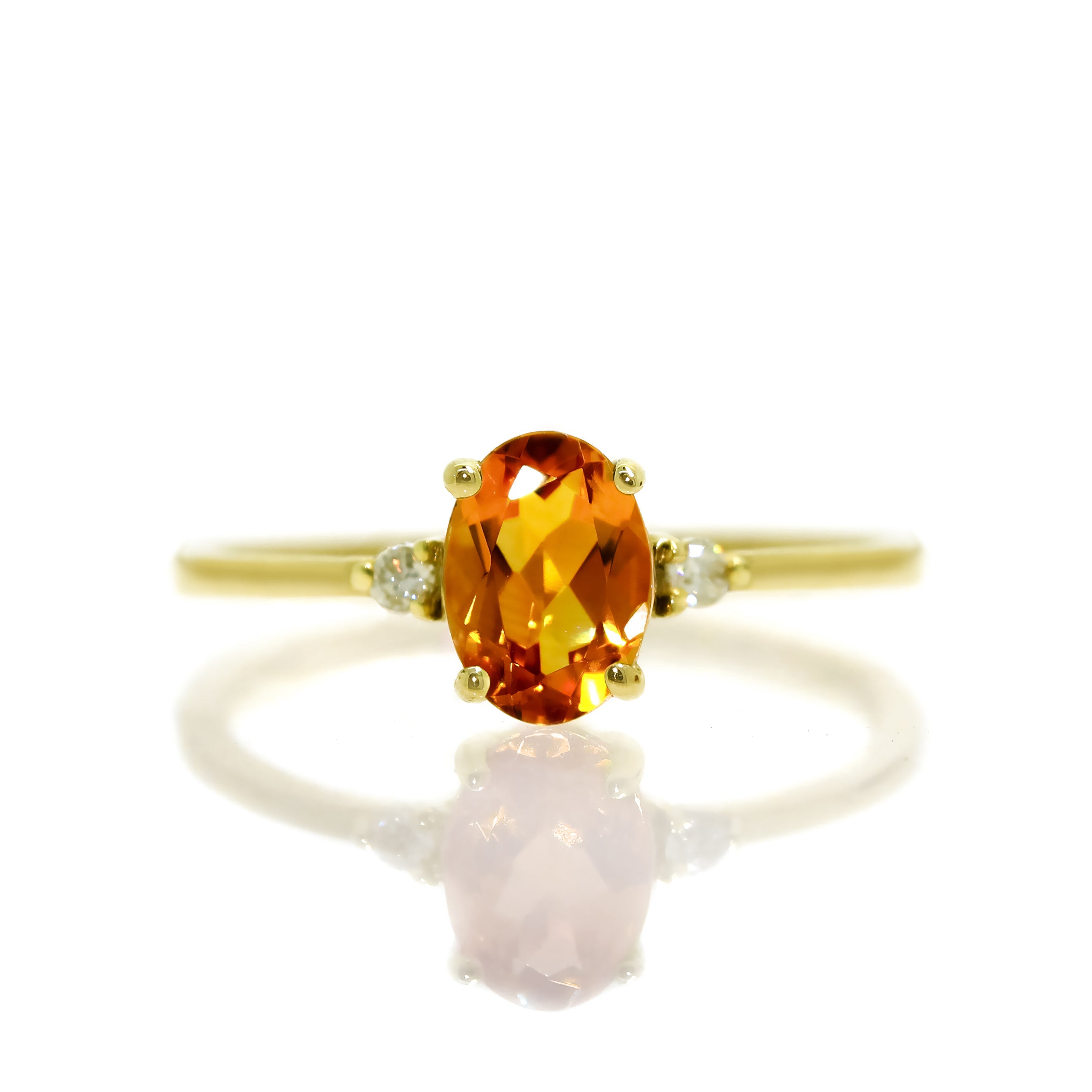 A product photo of a yellow gold citrine ring sitting against a white background. The yellow gold band is plain and smooth, and the centre oval-cut bright orange citrine stone is framed by a single white diamond on either side.