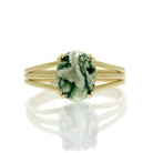 A product photo of a yellow gold moss agate ring sitting on a white background. The moss agate is impressively large, with the cabochon cut allowing the viewer to see every detail of its swirling, ink-like dendritic inclusions. The bandsplits into three prongs on either side of the stone.