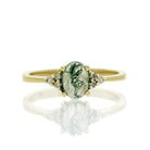 A product photo of a yellow gold ring with trio diamond detailing and a cabochon cut moss agate centre stone. The band is simple and smooth, with the cabochon cut of the moss agate allowing the viewer to see every detail of its swirling, ink-like dendritic inclusions.