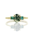 A product photo of a cabochon moss agate and mint blue tourmaline ring in 9 karat yellow gold on a white background. The cabochon cut of the moss agate allowing the viewer to see every detail of its swirling, ink-like dendritic inclusions, while the round faceted mint blue tourmaline adds a burst of colour.