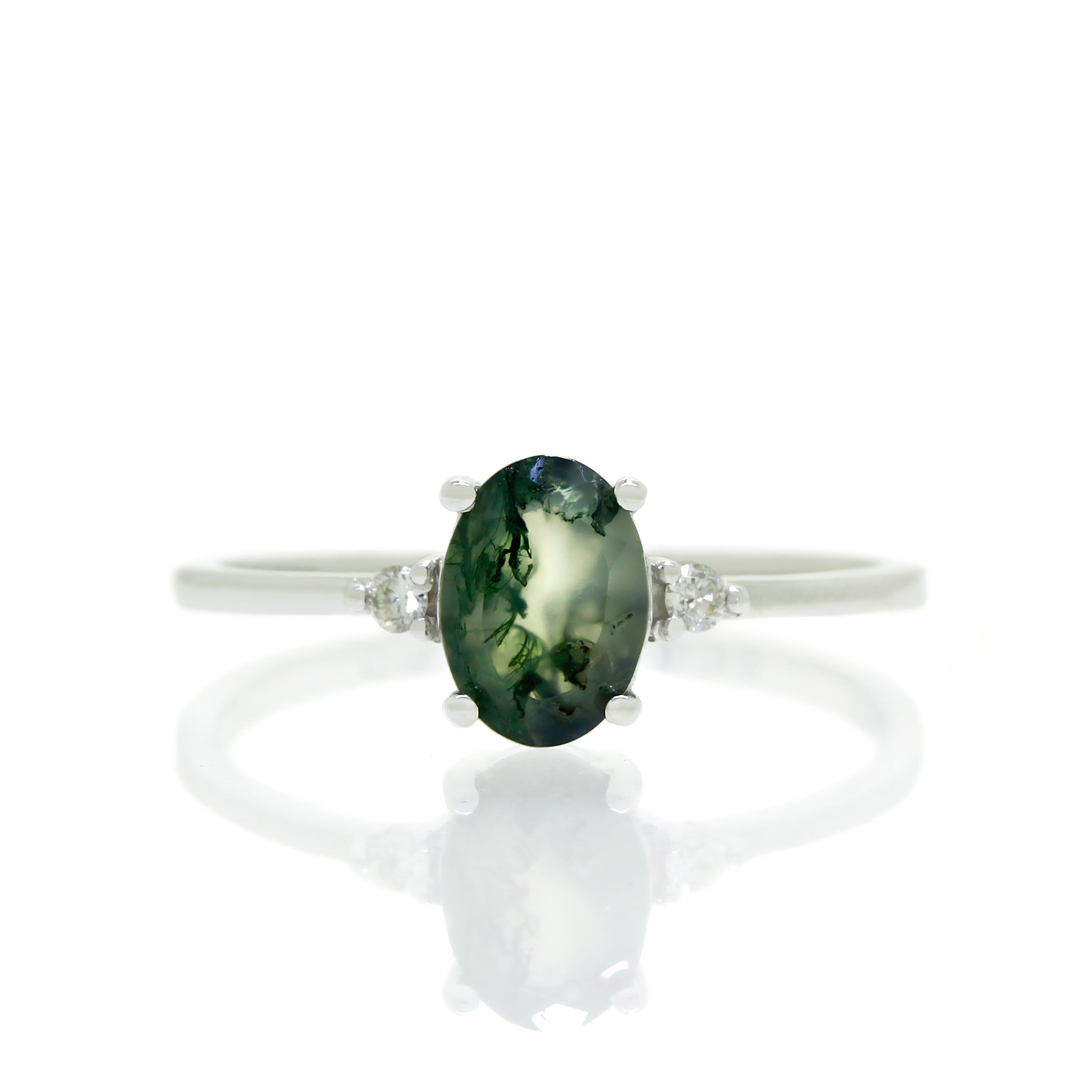 A product photo of a white gold moss agate ring sitting against a white background. The white gold band is plain and smooth, and the centre oval-cut deep green moss agate quartz stone is framed by a single white diamond on either side.