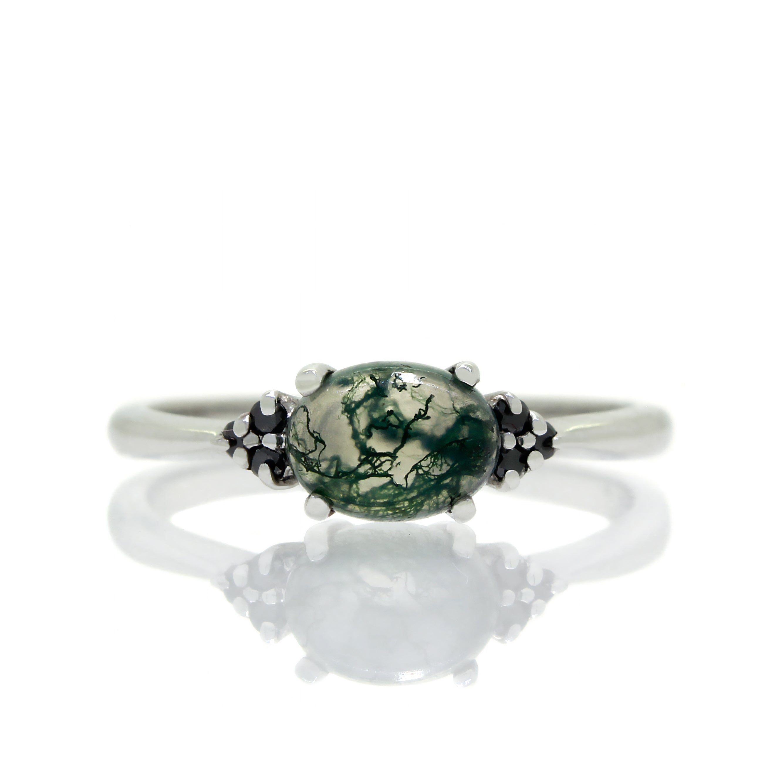 A product photo of a white gold moss agate and black diamond ring sitting against a white background. The white gold band is plain and smooth, and the horizontally-oriented cabochon moss agate centre stone is framed by a delicate trio of black diamonds on either side.