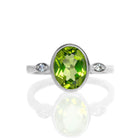 A product photo of a silver ring with a large bezel-set peridot centre stone sitting on a white background. The silver band is simple and smooth, connecting on either side of a vertically-oriented oval-cut peridot stone surrounded by a solid frame of silver. Before the band reaches the centre stone, it splits into two small horizontal silver oval frames on each side, each housing a small diamond.