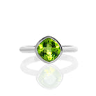 A product photo of a silver ring with a bezel-set peridot centre stone sitting on a white background. The silver band is simple and smooth, connecting on either side of a diagonally-oriented squared-cushion peridot stone surrounded by a solid frame of silver.