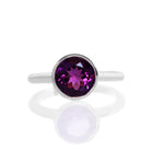 A product photo of a silver ring with a bezel-set amethyst centre stone sitting on a white background. The silver band is simple and smooth, connecting on either side of a large, statement-sized garnet stone surrounded by a solid frame of silver.