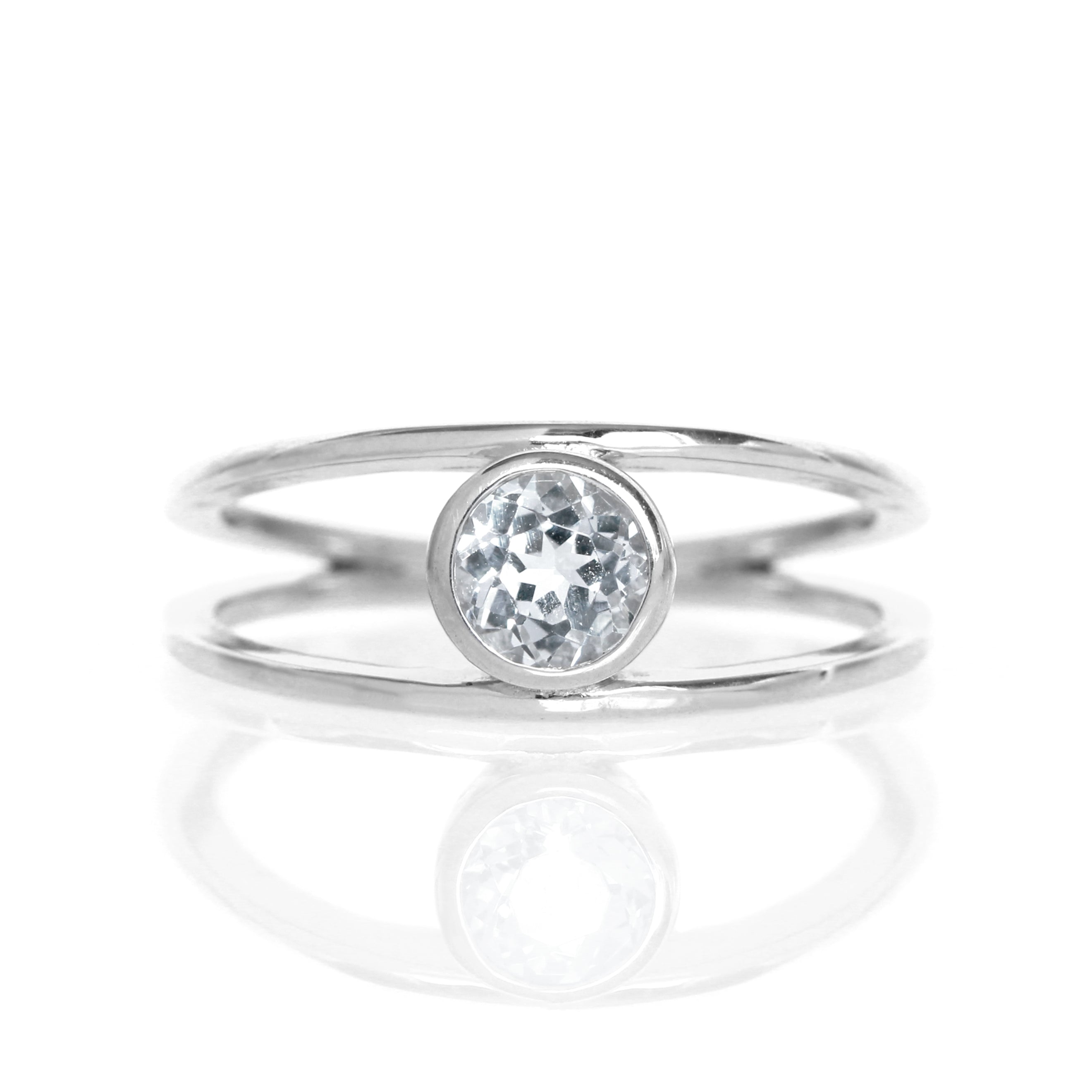 A product photo of a split-band silver ring with a bezel-set silver topaz centre stone sitting on a white background. The silver band splits at the base of the ring, separating to meet at the top and bottom of the bright white round-cut centre stone, framed in a thick layer of silver, holding it in place.