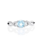 A product photo of an ornate silver ring with an aquamarine centre stone sitting on a white background. The silver band splits halfway along its length, becoming twisting and serpentine in appearance before meeting on either side of the dazzling blue 5mm stone, which is held in place by 4 silver claws.