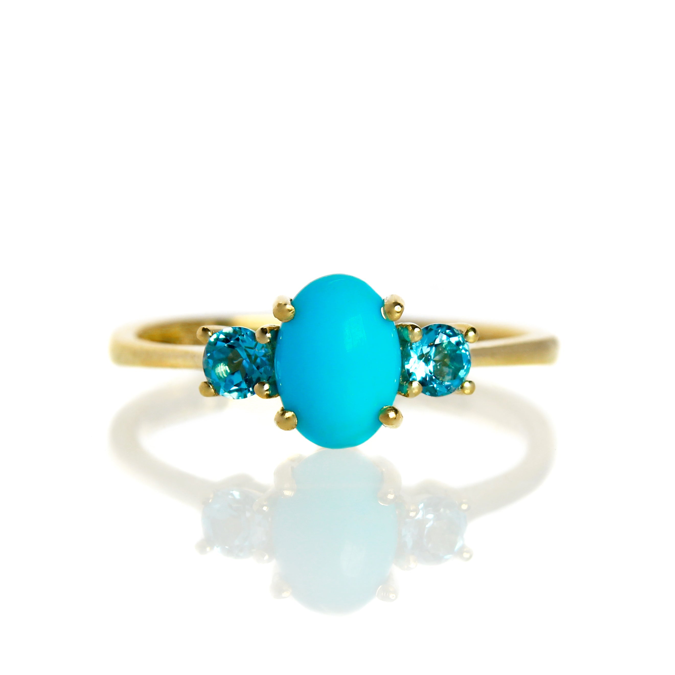 A product photo of a cabochon Sleeping Beauty turquoise and blue topaz ring in 9 karat yellow gold on a white background. The 7x5mm turquoise stone is perfectly opaque and unblemished, and is hugged on either side by light blue 3mm round blue topaz jewels.