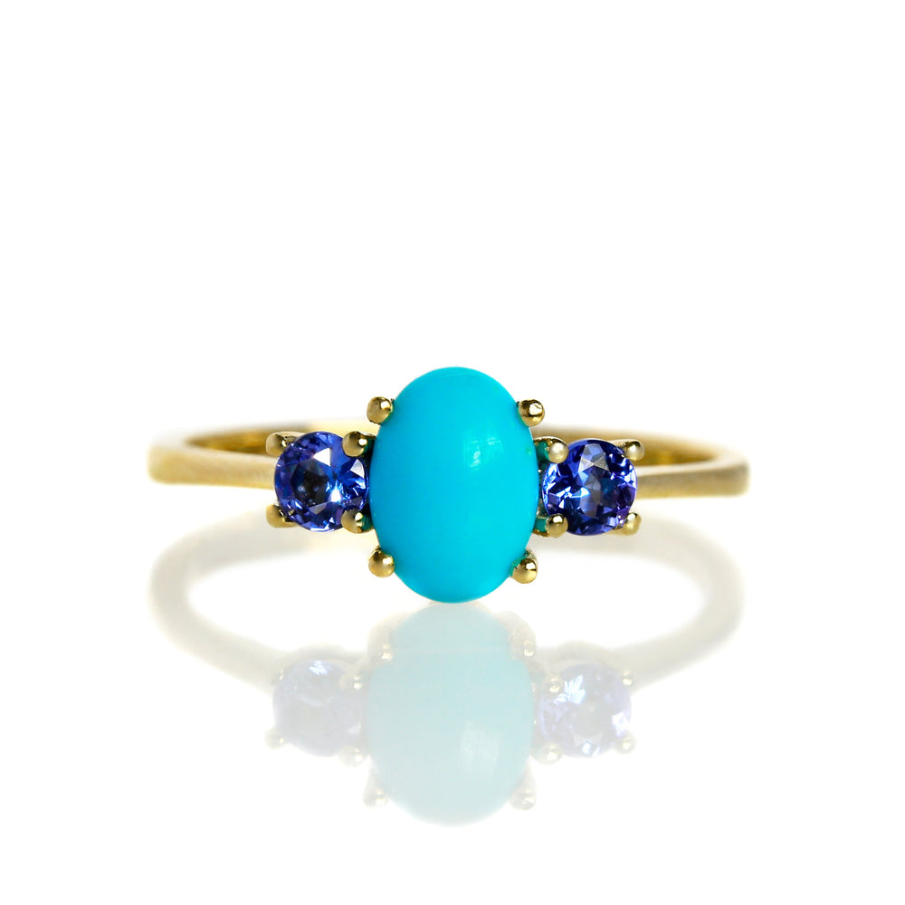 A product photo of a cabochon Sleeping Beauty turquoise and tanzanite birthstone ring in 9 karat yellow gold on a white background. The 7x5mm turquoise stone is perfectly opaque and unblemished, and is hugged on either side by light blue 3mm round blue topaz jewels.