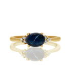 A product photo of a yellow gold cabochon star sapphire and diamond ring sitting against a white background. The yellow gold band is plain and smooth, and the horizontally-oriented oval cabochon cut stone is framed by a delicate trio of white diamonds on either side.