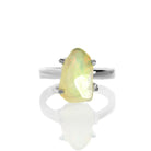 A product photo of a boho style statement ring on a white background. The ring is made up of a thick, flat sterling silver band, and a rough-cut rainbow opal gemstone. The stone is large and reflects a multitude of bright colours in irregular shapes.