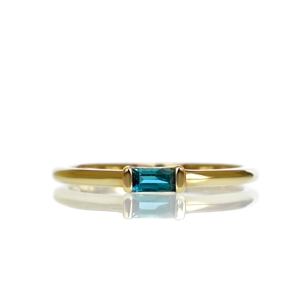 A product photo of a solid 9 karat yellow gold london blue topaz ring on a white background. The slim golden band is comfortably rounded, and the 4x2mm rectangular deep ocean blue topaz stone is held horizontally in place by one squared setting on either side.