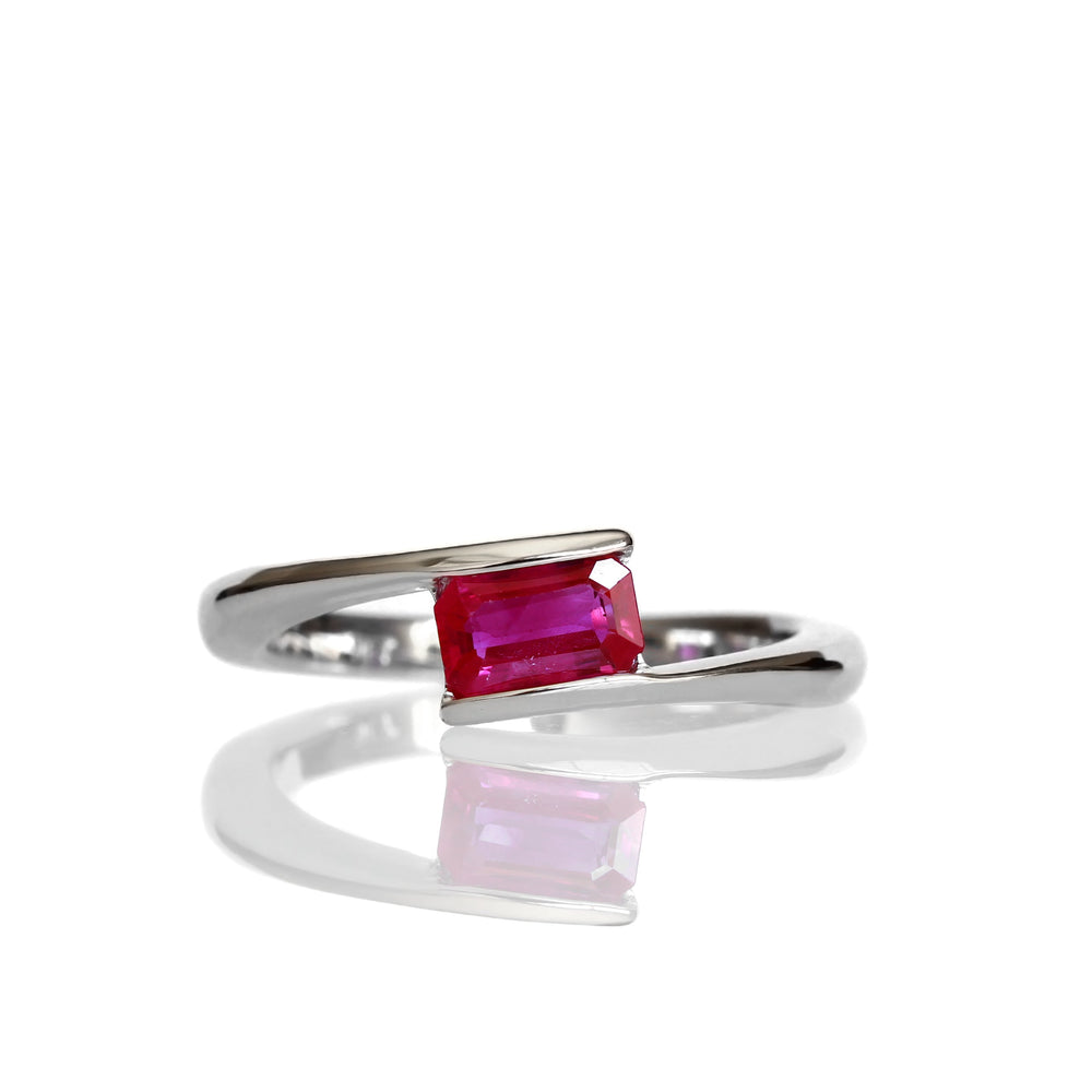 A product photo of an asymmetric ruby ring in 9 karat white gold on a white background. The smooth golden band is split, one side curving up and the other down to meet at the top and bottom of the horizontally-oriented pink ruby stone in an asymmetrical fashion.